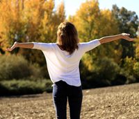 Frau mit ausgebreiteten Armen steht vor einem Herbstwald, gekleidet in ein weißes Shirt und dunkle Hose, symbolisiert Freiheit und Balance.