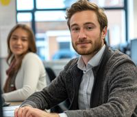 Junger Mann im Büro sitzt am Schreibtisch, lächelnd in die Kamera, trägt ein graues Tweed-Sakko und ein hellblaues Hemd. Im Hintergrund eine unscharf fokussierte junge Frau an einem weiteren Arbeitsplatz.