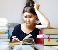 Besorgte junge Frau mit Hochsteckfrisur sitzt an einem Tisch, umgeben von Bücherstapeln, und liest ein Buch. Sie fasst sich mit einer Hand an den Kopf, was Verwirrung oder Stress andeutet.